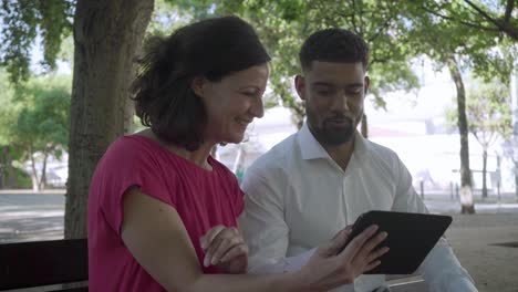 Cheerful-multiracial-couple-sitting-on-bench-and-using-tablet.